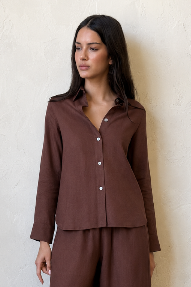 Woman wearing a brown linen shirt and pant standing against a white wall.
