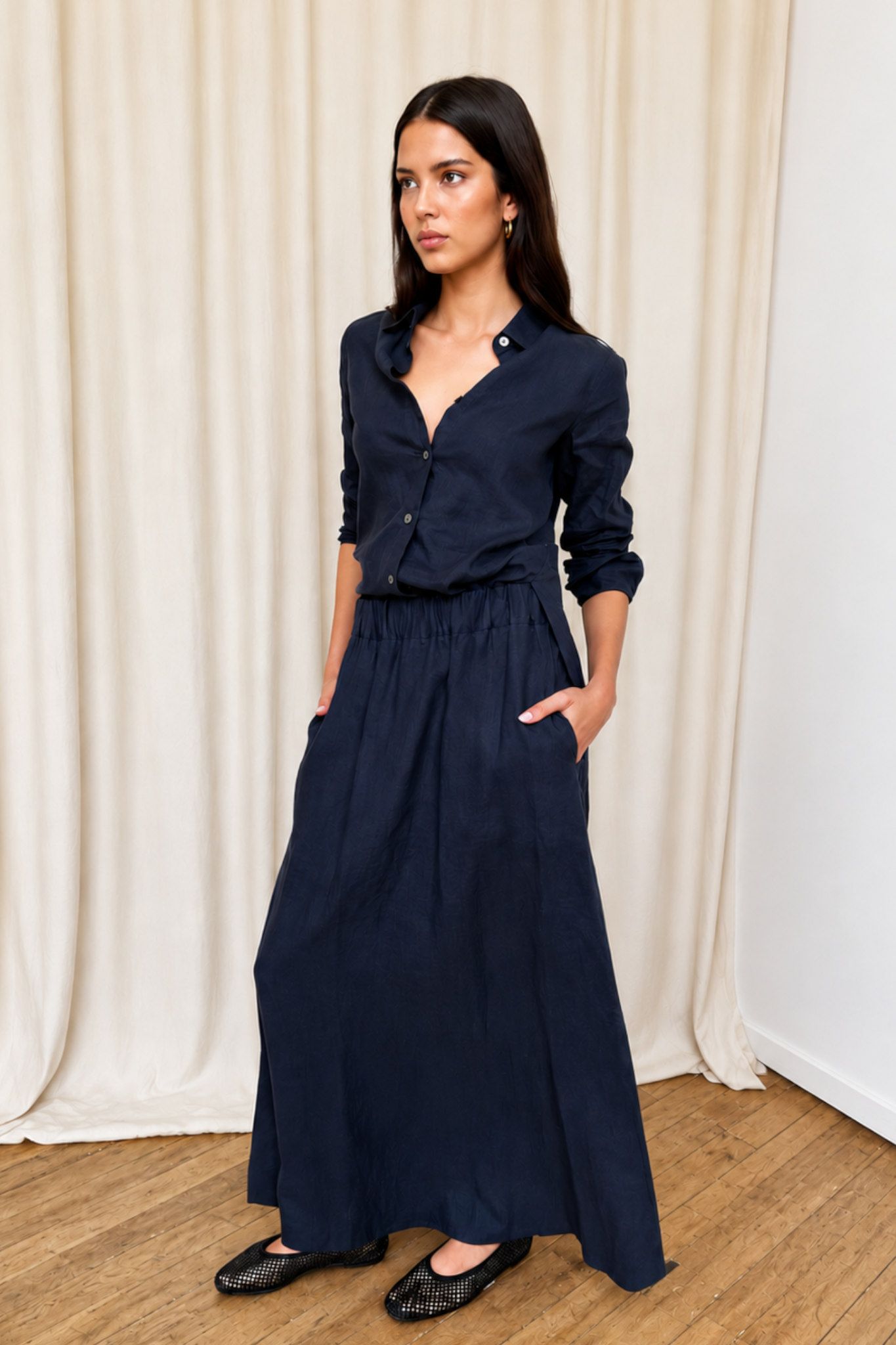 Woman wearing a navy blue linen shirt and a navy blue linen maxi skirt against a white curtain background